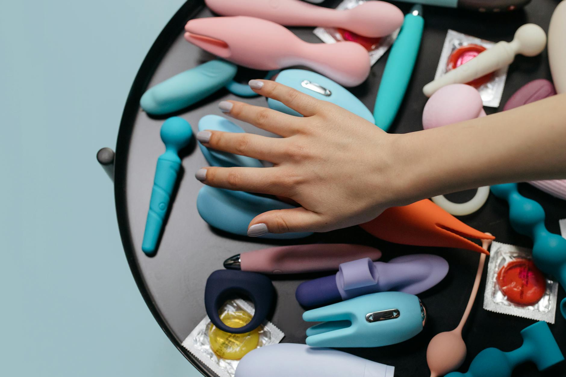 A hand reaching over a variety of colorful clitoral vibrators arranged on a table, showing different options for sensory exploration