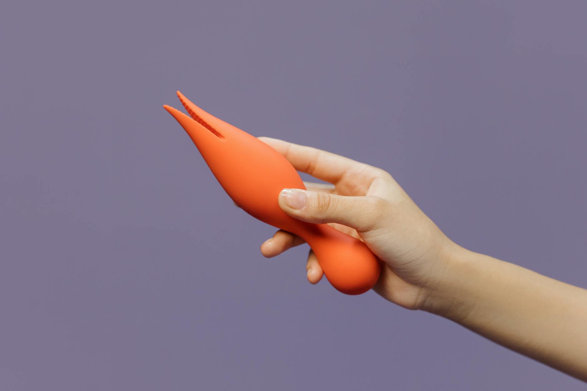 A person's hand holding an orange vibrator against a minimalistic purple backdrop